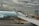 A plane from Air Canada is parked at an airport gate with a cloudy sky overhead. The airplane's wing is prominent in the foreground. Ground support vehicles and equipment are visible around the aircraft, along with a jet bridge leading to the plane. A digital sign with a maple leaf logo is displayed near the gate.