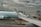 A plane from Air Canada is parked at an airport gate with a cloudy sky overhead. The airplane's wing is prominent in the foreground. Ground support vehicles and equipment are visible around the aircraft, along with a jet bridge leading to the plane. A digital sign with a maple leaf logo is displayed near the gate.