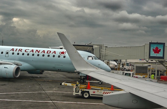A plane from Air Canada is parked at an airport gate with a cloudy sky overhead. The airplane's wing is prominent in the foreground. Ground support vehicles and equipment are visible around the aircraft, along with a jet bridge leading to the plane. A digital sign with a maple leaf logo is displayed near the gate.