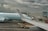 A plane from Air Canada is parked at an airport gate with a cloudy sky overhead. The airplane's wing is prominent in the foreground. Ground support vehicles and equipment are visible around the aircraft, along with a jet bridge leading to the plane. A digital sign with a maple leaf logo is displayed near the gate.