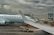 A plane from Air Canada is parked at an airport gate with a cloudy sky overhead. The airplane's wing is prominent in the foreground. Ground support vehicles and equipment are visible around the aircraft, along with a jet bridge leading to the plane. A digital sign with a maple leaf logo is displayed near the gate.