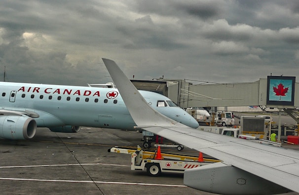 A plane from Air Canada is parked at an airport gate with a cloudy sky overhead. The airplane's wing is prominent in the foreground. Ground support vehicles and equipment are visible around the aircraft, along with a jet bridge leading to the plane. A digital sign with a maple leaf logo is displayed near the gate.