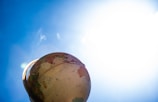 A globe is positioned against a bright blue sky, slightly tilted to reveal parts of Africa and South America. The sunlight creates a lens flare effect on the left, with the top part of the globe illuminated.