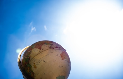A globe is positioned against a bright blue sky, slightly tilted to reveal parts of Africa and South America. The sunlight creates a lens flare effect on the left, with the top part of the globe illuminated.