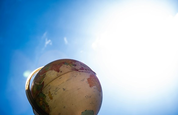 A globe is positioned against a bright blue sky, slightly tilted to reveal parts of Africa and South America. The sunlight creates a lens flare effect on the left, with the top part of the globe illuminated.