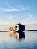 A large cargo ship is carrying multiple stacked shipping containers across a calm body of water under a clear blue sky with some wispy clouds. The peaceful reflections in the water highlight the tranquil setting as the ship moves toward the horizon.