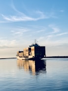 A large cargo ship is carrying multiple stacked shipping containers across a calm body of water under a clear blue sky with some wispy clouds. The peaceful reflections in the water highlight the tranquil setting as the ship moves toward the horizon.