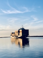 A large cargo ship is carrying multiple stacked shipping containers across a calm body of water under a clear blue sky with some wispy clouds. The peaceful reflections in the water highlight the tranquil setting as the ship moves toward the horizon.