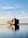 A large cargo ship is carrying multiple stacked shipping containers across a calm body of water under a clear blue sky with some wispy clouds. The peaceful reflections in the water highlight the tranquil setting as the ship moves toward the horizon.