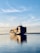 A large cargo ship is carrying multiple stacked shipping containers across a calm body of water under a clear blue sky with some wispy clouds. The peaceful reflections in the water highlight the tranquil setting as the ship moves toward the horizon.