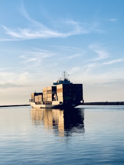 A large cargo ship is carrying multiple stacked shipping containers across a calm body of water under a clear blue sky with some wispy clouds. The peaceful reflections in the water highlight the tranquil setting as the ship moves toward the horizon.