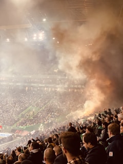A crowded sports stadium filled with spectators, with smoke billowing in the air creating a hazy atmosphere. Bright stadium lights illuminate the scene, and people can be seen clapping or raising their hands, suggesting an intense moment in the event.