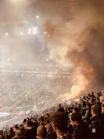 A crowded sports stadium filled with spectators, with smoke billowing in the air creating a hazy atmosphere. Bright stadium lights illuminate the scene, and people can be seen clapping or raising their hands, suggesting an intense moment in the event.