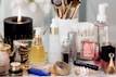 A collection of beauty and skincare products is arranged on a surface, including bottles of oils and serums, cosmetics like lipstick and blush, makeup brushes in a holder, and a lit black candle adding ambiance. The setup is accompanied by cotton swabs, emphasizing a self-care or beauty routine theme.
