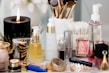 A collection of beauty and skincare products is arranged on a surface, including bottles of oils and serums, cosmetics like lipstick and blush, makeup brushes in a holder, and a lit black candle adding ambiance. The setup is accompanied by cotton swabs, emphasizing a self-care or beauty routine theme.