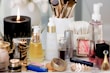 A collection of beauty and skincare products is arranged on a surface, including bottles of oils and serums, cosmetics like lipstick and blush, makeup brushes in a holder, and a lit black candle adding ambiance. The setup is accompanied by cotton swabs, emphasizing a self-care or beauty routine theme.