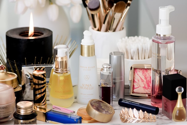 A collection of beauty and skincare products is arranged on a surface, including bottles of oils and serums, cosmetics like lipstick and blush, makeup brushes in a holder, and a lit black candle adding ambiance. The setup is accompanied by cotton swabs, emphasizing a self-care or beauty routine theme.
