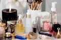 A collection of beauty and skincare products is arranged on a surface, including bottles of oils and serums, cosmetics like lipstick and blush, makeup brushes in a holder, and a lit black candle adding ambiance. The setup is accompanied by cotton swabs, emphasizing a self-care or beauty routine theme.