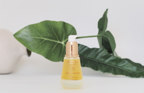 A bottle of skin care product labeled 'Sonia Roselli Water Oil Skin Prep' stands on a surface. Behind it, large green leaves and a white vase partially visible to the left create a natural backdrop. The product has a white and gold pump dispenser, with the body of the bottle in translucent ivory, giving a luxurious and fresh appearance.