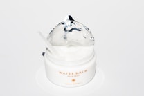 A white jar of Water Balm Skin Prep with a silver foil lid partially opened. It contains a creamy substance, and a small spatula rests inside the jar. The product label is printed in gold lettering.