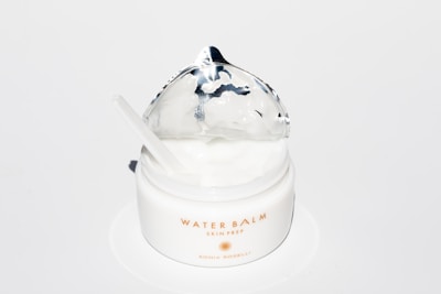 A white jar of Water Balm Skin Prep with a silver foil lid partially opened. It contains a creamy substance, and a small spatula rests inside the jar. The product label is printed in gold lettering.