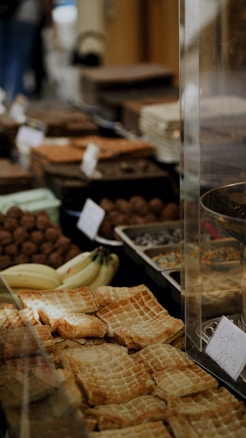 A variety of pastries and baked goods are displayed on a table, including flaky pastries and loaves. There are also bananas and various other pieces of bread and confections, with some items labeled with price tags.