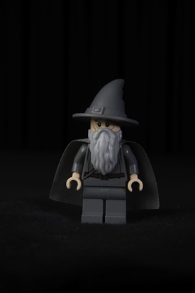 A LEGO minifigure dressed as a wizard, featuring a tall pointed hat, a long gray beard, and a dark cape. The figure stands against a black background, emphasizing its detailed design and muted color scheme.