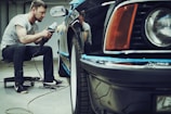 A man is carefully polishing a black car with a buffer in a garage setting. The focus is on his concentration and the shiny surface of the vehicle. He is seated on a small rolling stool to comfortably reach the side of the car.
