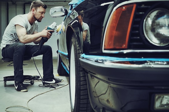 A man is carefully polishing a black car with a buffer in a garage setting. The focus is on his concentration and the shiny surface of the vehicle. He is seated on a small rolling stool to comfortably reach the side of the car.