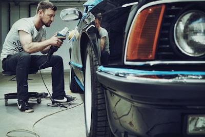 A man is carefully polishing a black car with a buffer in a garage setting. The focus is on his concentration and the shiny surface of the vehicle. He is seated on a small rolling stool to comfortably reach the side of the car.