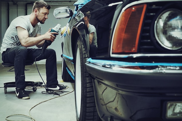 A man is carefully polishing a black car with a buffer in a garage setting. The focus is on his concentration and the shiny surface of the vehicle. He is seated on a small rolling stool to comfortably reach the side of the car.
