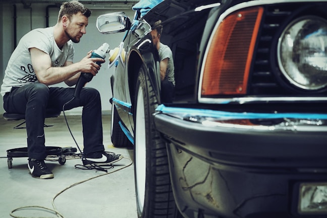 A man is carefully polishing a black car with a buffer in a garage setting. The focus is on his concentration and the shiny surface of the vehicle. He is seated on a small rolling stool to comfortably reach the side of the car.