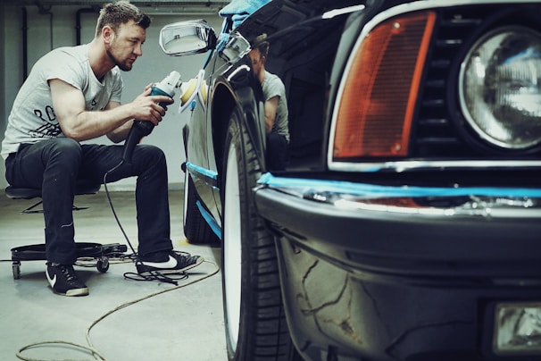 A man is carefully polishing a black car with a buffer in a garage setting. The focus is on his concentration and the shiny surface of the vehicle. He is seated on a small rolling stool to comfortably reach the side of the car.