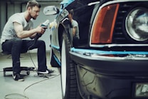 A man is carefully polishing a black car with a buffer in a garage setting. The focus is on his concentration and the shiny surface of the vehicle. He is seated on a small rolling stool to comfortably reach the side of the car.
