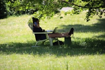 A person is relaxing on a lawn chair in a grassy area, partially shaded by tree branches. They are using a smartphone and have a laptop open on a small table in front of them. The scene suggests leisure or remote work in an outdoor setting.