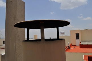 A rooftop scene with a prominent cylindrical structure in the foreground featuring a flat, overhanging lid. The background includes white buildings, with one having a bright red surface. A solar water heater and a tall piece of equipment are visible against a partly cloudy blue sky.
