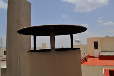 A rooftop scene with a prominent cylindrical structure in the foreground featuring a flat, overhanging lid. The background includes white buildings, with one having a bright red surface. A solar water heater and a tall piece of equipment are visible against a partly cloudy blue sky.