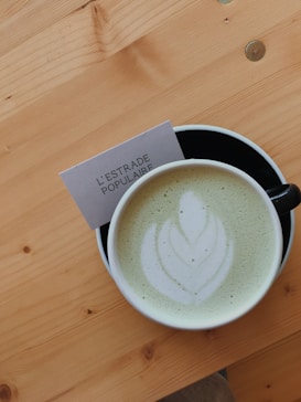 A cup of matcha latte with an intricate leaf pattern created in the foam, placed on a light wooden table. Beside the cup, a business card with the text 'L'ESTRADE POPULAIRE' is partially visible.