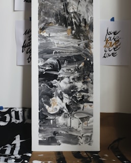 An abstract painting dominated by black, white, and gray tones with some hints of gold. The artwork features fluid, swirling patterns with a mix of textures. It is surrounded by sheets of paper taped to the wall with the word 'love' written repeatedly in varying styles and colors.