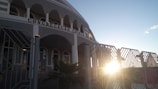 A building with arched windows and a facade that has the words 'Congregacao Crista no Brasil' displayed prominently. The architectural design includes multiple columns and a gated entrance. The sun is setting or rising, creating a bright glow and casting shadows.