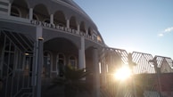 A building with arched windows and a facade that has the words 'Congregacao Crista no Brasil' displayed prominently. The architectural design includes multiple columns and a gated entrance. The sun is setting or rising, creating a bright glow and casting shadows.