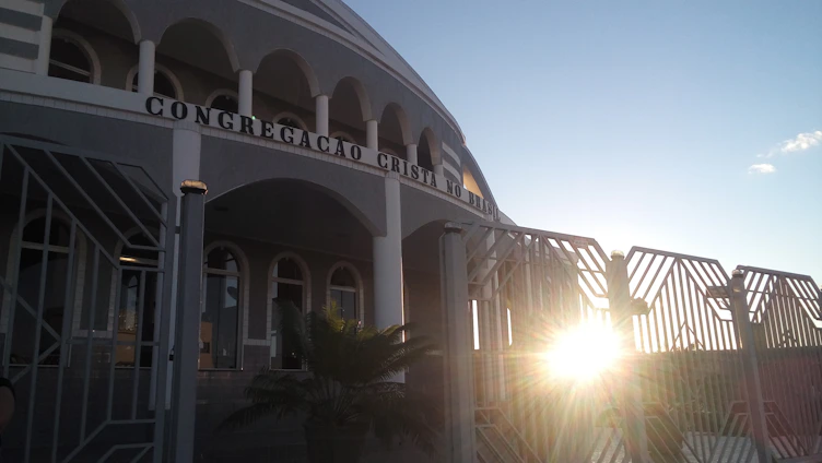 A building with arched windows and a facade that has the words 'Congregacao Crista no Brasil' displayed prominently. The architectural design includes multiple columns and a gated entrance. The sun is setting or rising, creating a bright glow and casting shadows.