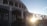 A building with arched windows and a facade that has the words 'Congregacao Crista no Brasil' displayed prominently. The architectural design includes multiple columns and a gated entrance. The sun is setting or rising, creating a bright glow and casting shadows.