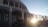 A building with arched windows and a facade that has the words 'Congregacao Crista no Brasil' displayed prominently. The architectural design includes multiple columns and a gated entrance. The sun is setting or rising, creating a bright glow and casting shadows.