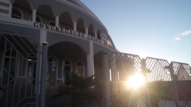 A building with arched windows and a facade that has the words 'Congregacao Crista no Brasil' displayed prominently. The architectural design includes multiple columns and a gated entrance. The sun is setting or rising, creating a bright glow and casting shadows.
