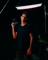 A person stands confidently holding a camera with studio lighting from above, surrounded by a dark backdrop and a few visible green plants on the side. The lighting creates a dramatic contrast, highlighting the subject.