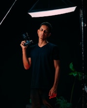 A person stands confidently holding a camera with studio lighting from above, surrounded by a dark backdrop and a few visible green plants on the side. The lighting creates a dramatic contrast, highlighting the subject.