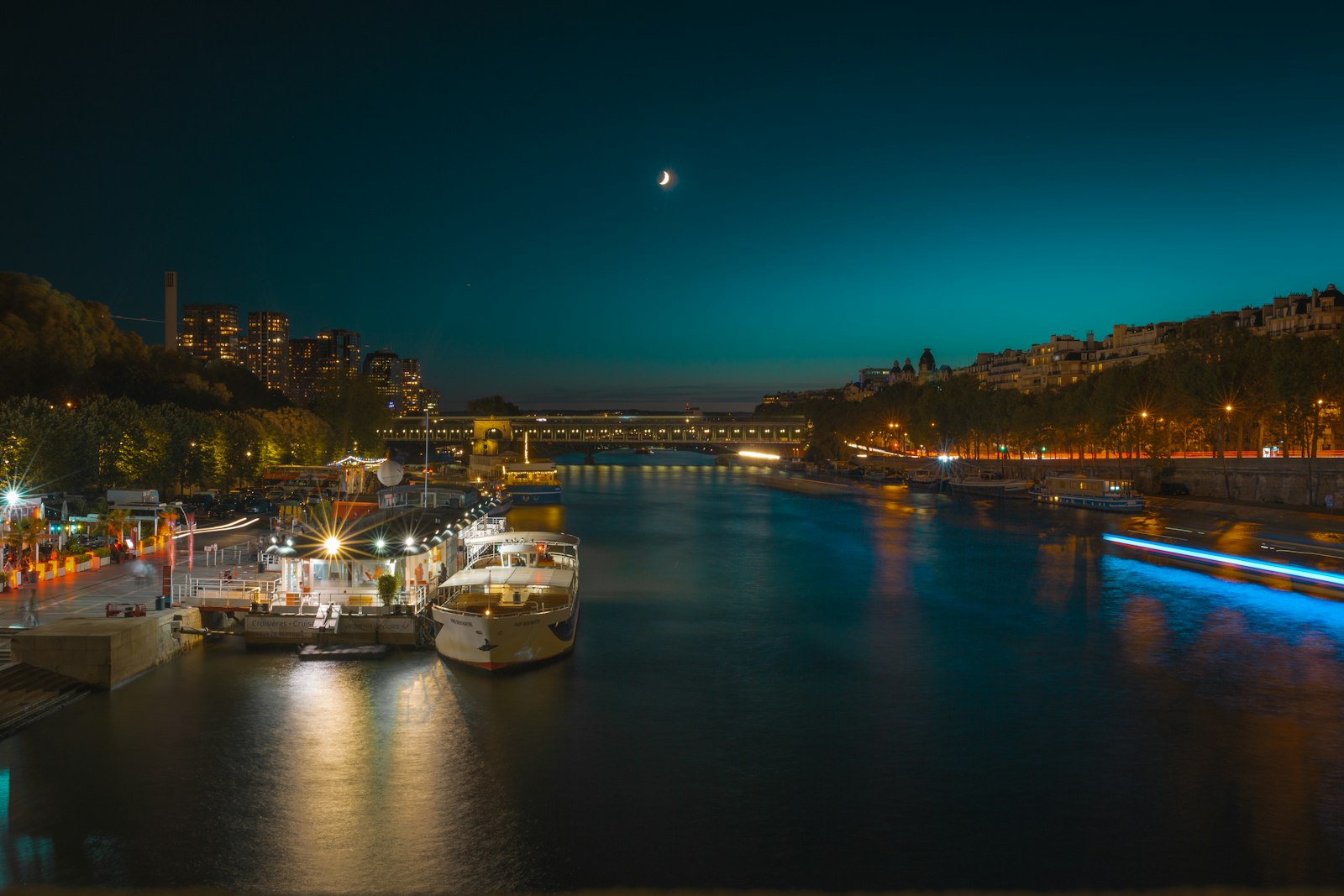 Boats moored on the dark Seine at night under a crescent moon, with lit bridges and riverbanks in the distance