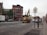 A city street with construction equipment, including a large excavator from CAT parked in the middle of the road. The street is lined with buildings featuring various architectural designs, including a reddish-brown brick building. There are cars parked and in motion, along with pedestrians walking on the sidewalks. Traffic cones and signs are visible, indicating ongoing roadwork.