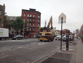 A city street with construction equipment, including a large excavator from CAT parked in the middle of the road. The street is lined with buildings featuring various architectural designs, including a reddish-brown brick building. There are cars parked and in motion, along with pedestrians walking on the sidewalks. Traffic cones and signs are visible, indicating ongoing roadwork.